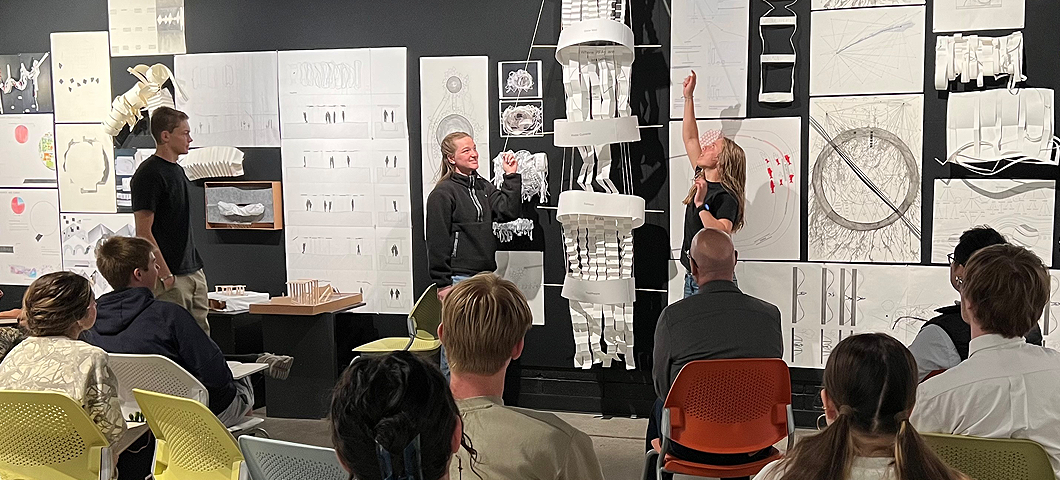 Two students present interactive paper sculpture to Faculty Mike Everts and James Park while the Lower Gallery audience looks on.